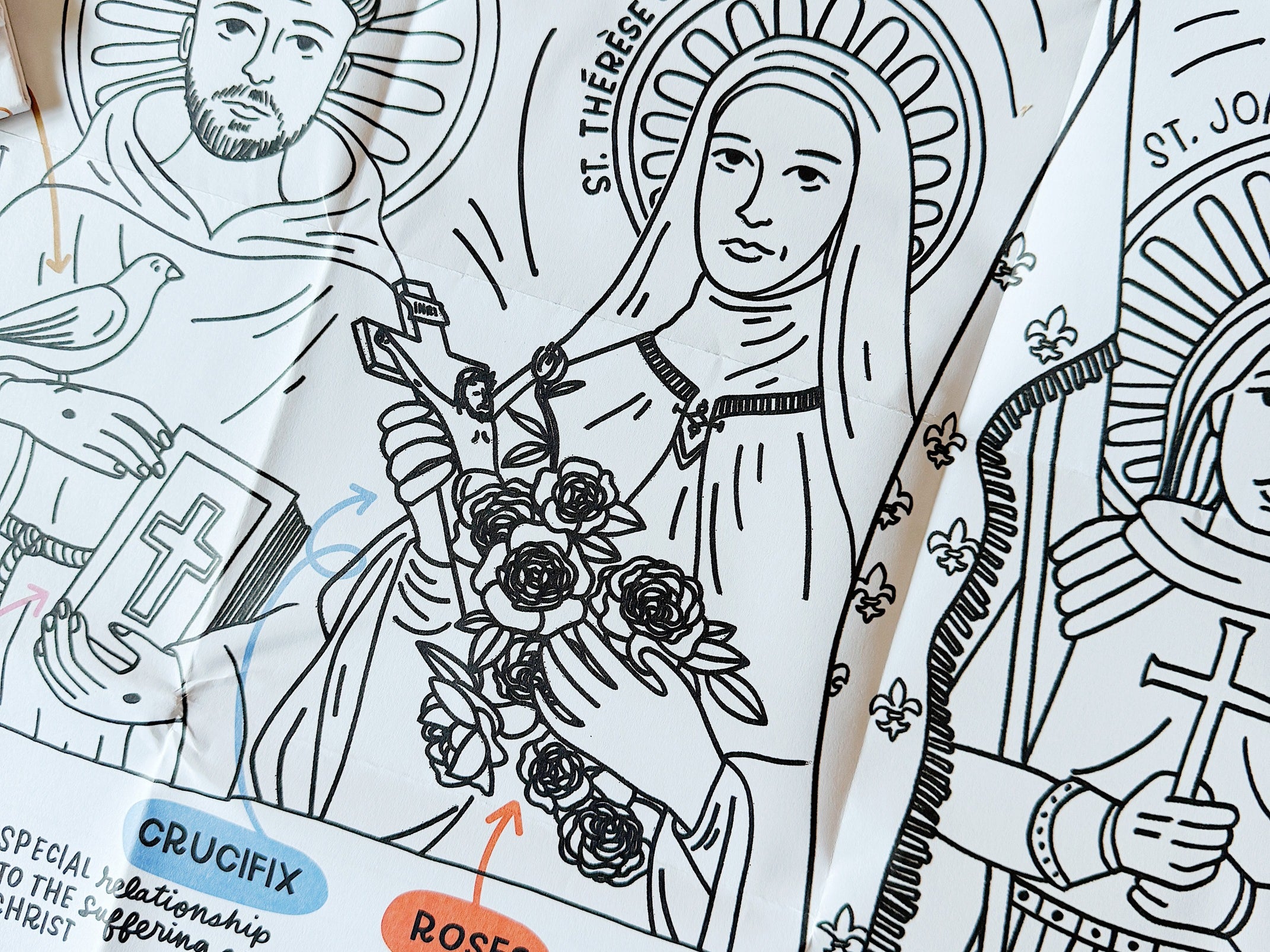 Close-up of a folded black-and-white iconography coloring sheet showing saints (St. Francis of Assisi, St. Thérèse of Lisieux) with labeled symbols like halo, crown, crucifix, roses, palm, and open Bible explaining their meanings.