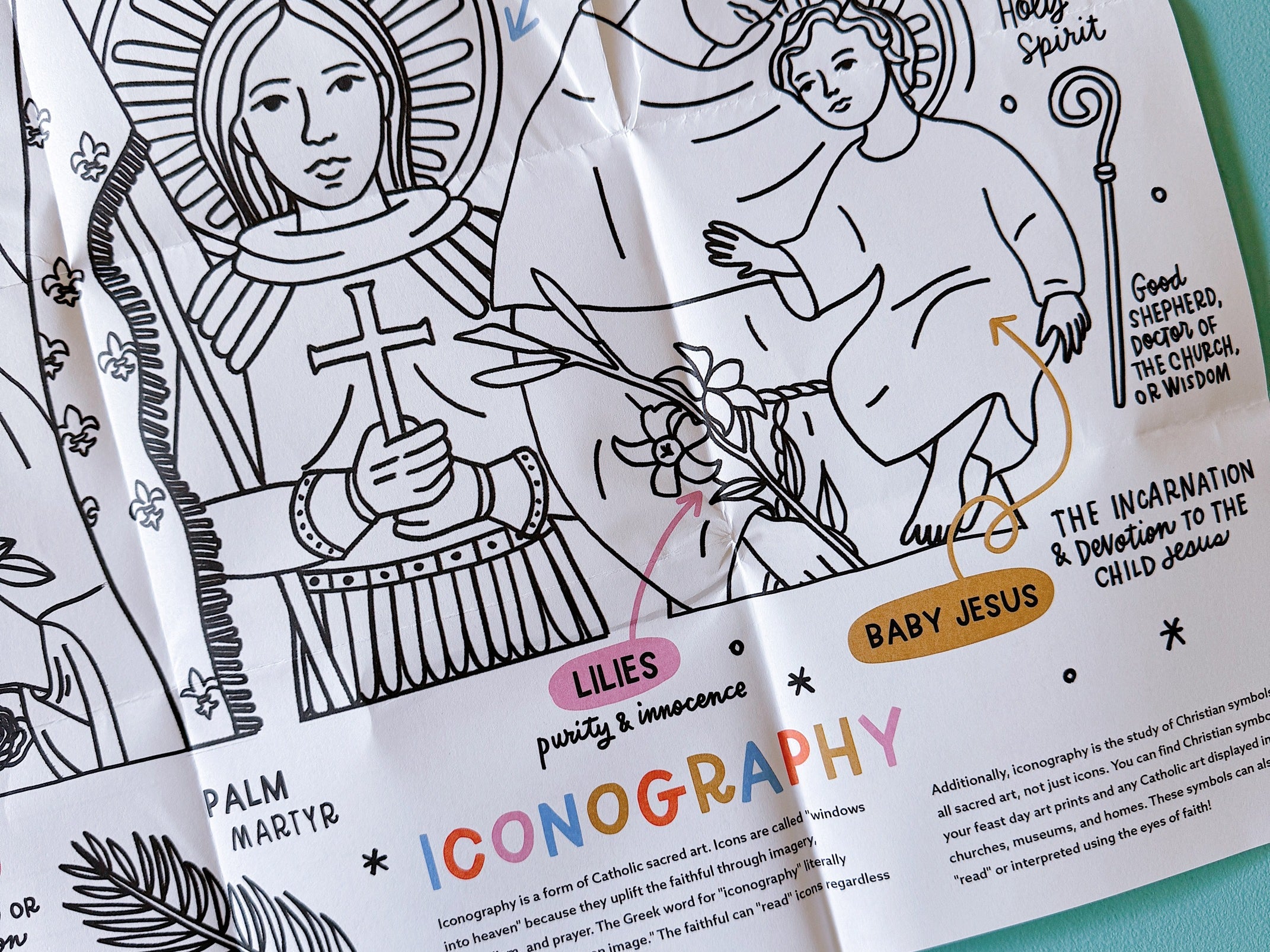 Folded black-and-white coloring sheet showing St. Joan of Arc and St. Anthony of Padua with Baby Jesus, labeled with symbols (armor, lilies, dove, palm) and a short explanation of Catholic iconography.